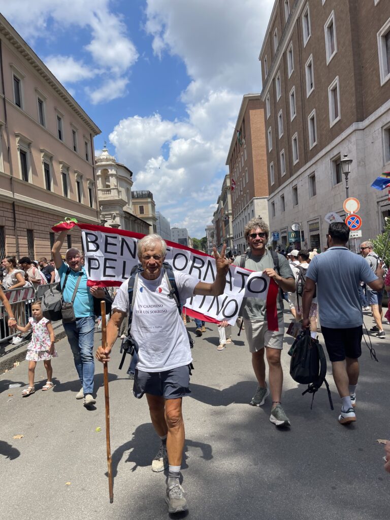 Stefano Barbero “Un uomo anziano con capelli bianchi, vestito con maglietta bianca e pantaloncini, cammina sorridente in una strada affollata, tenendo un bastone da cammino. Dietro di lui, alcune persone sorreggono uno striscione con la scritta ‘BENTORNATO PELLEGRINO’.